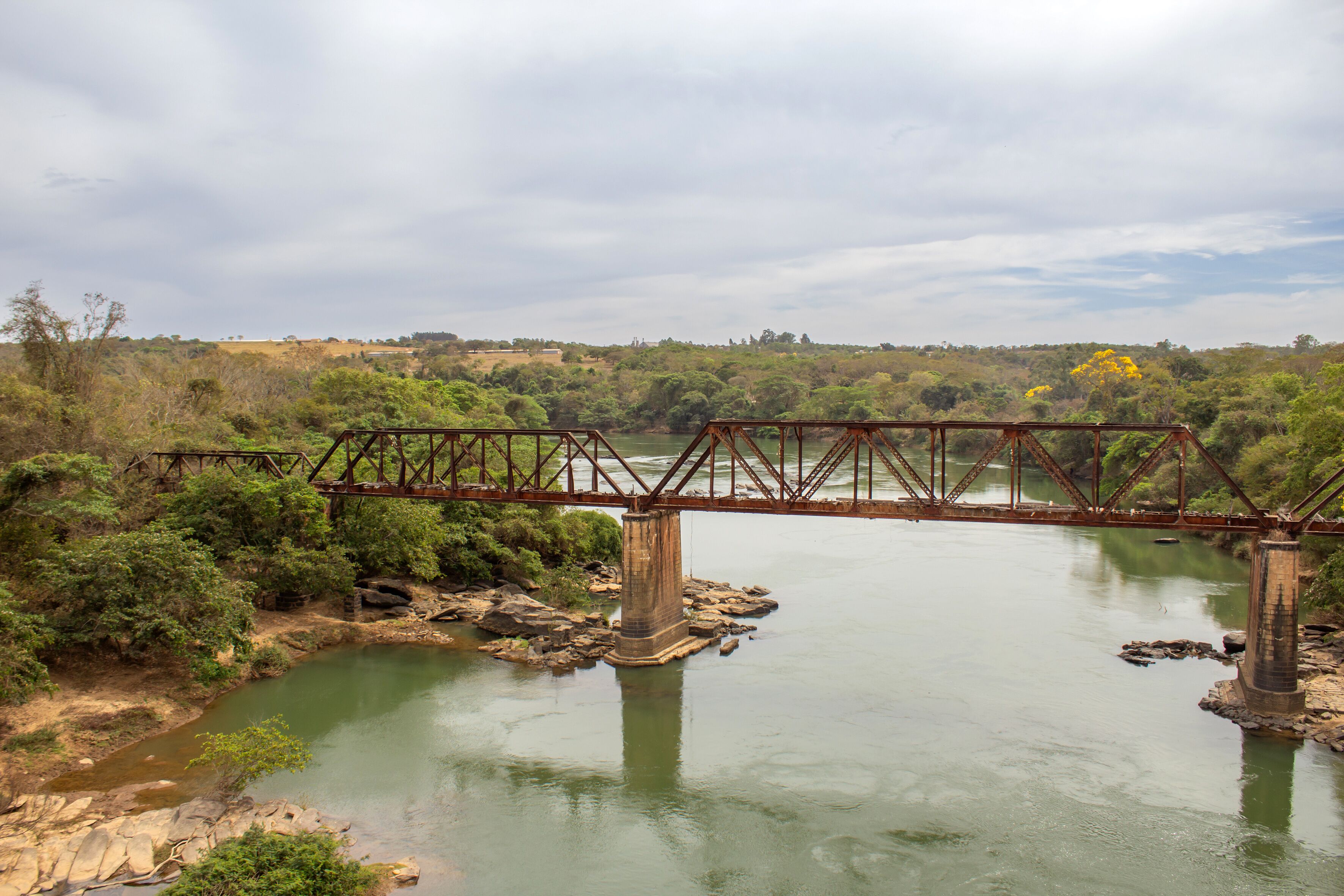 A rusted metal bridge over a calm river surrounded by dense greenery and hills.