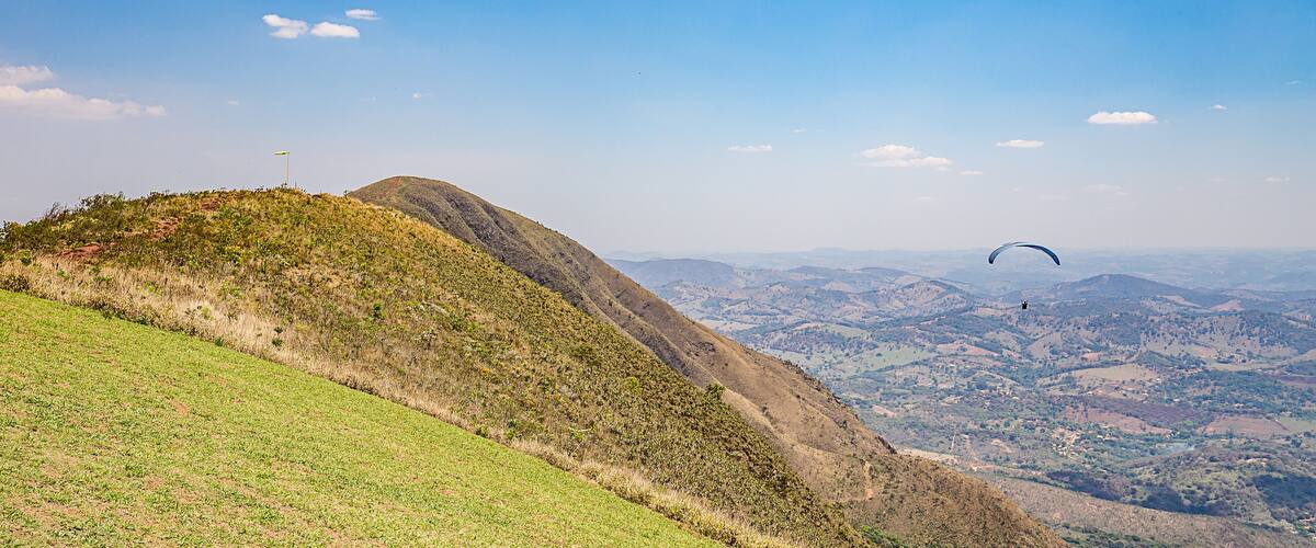 A grassy hill with a flag, a valley, and a paraglider in the distance.