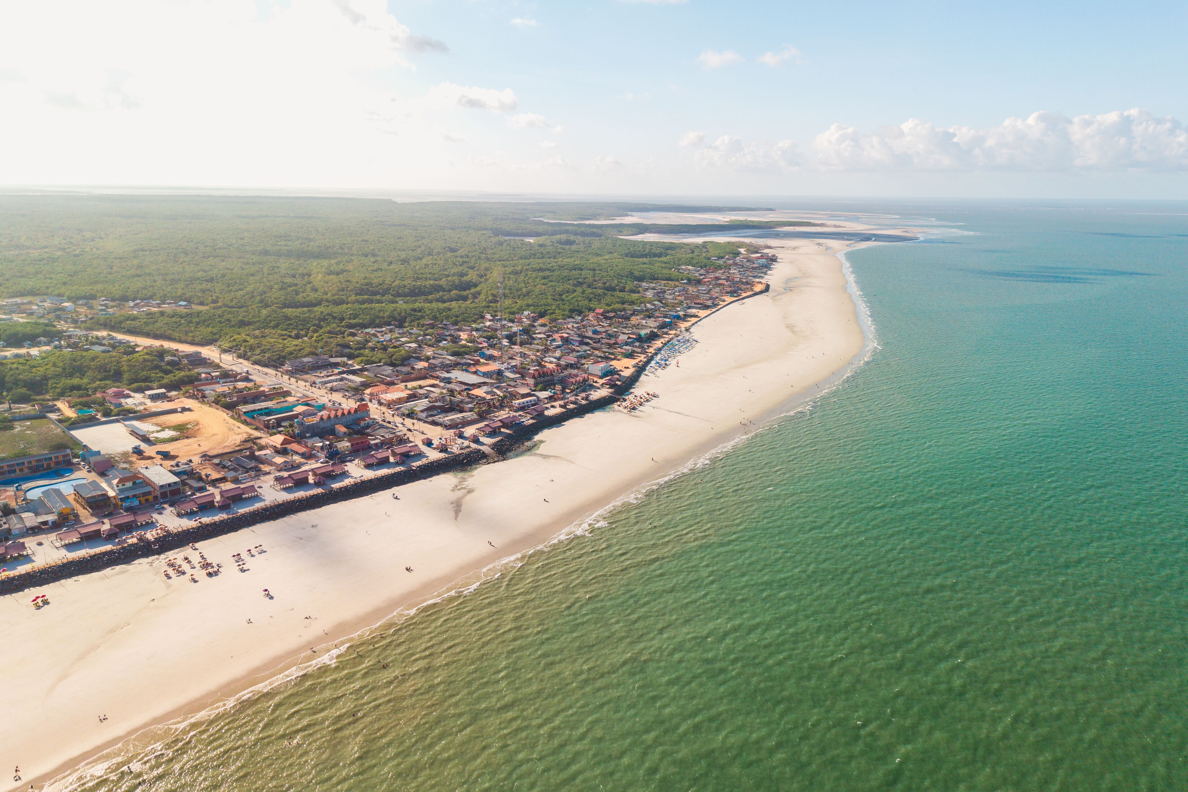 An aerial view of a coastal town with a long beach, clear turquoise waters, and dense green vegetation inland.