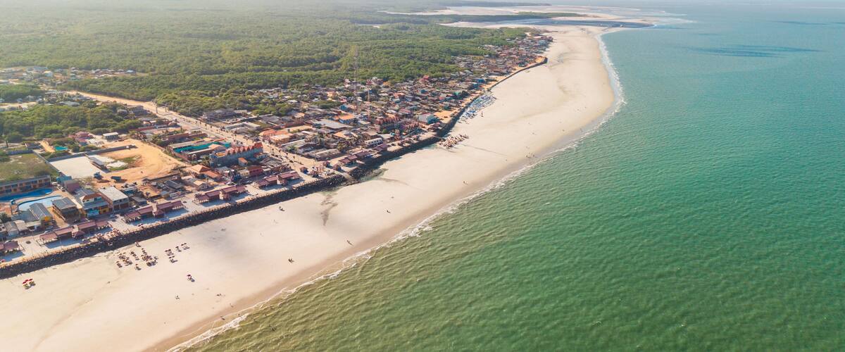 An aerial view of a coastal town with a long beach, clear turquoise waters, and dense green vegetation inland.