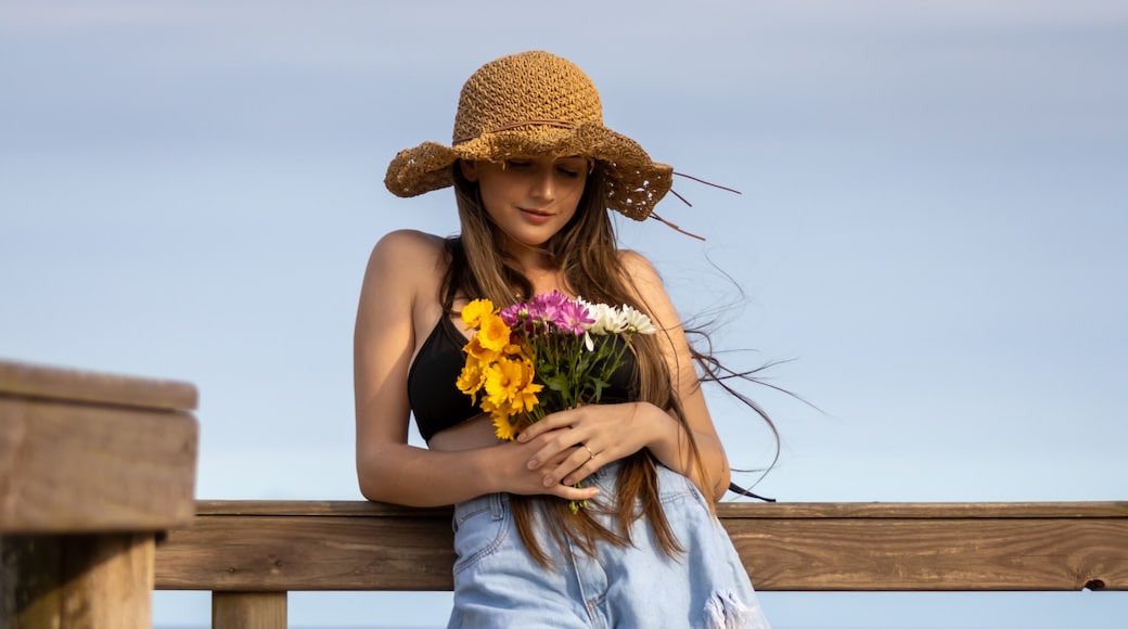 Balneário Gaivota, Santa Catarina, Brazil, November 2020. Model in front of the sea holding beautiful colorful flowers with a wavy brown hat illuminated by the sunlight.