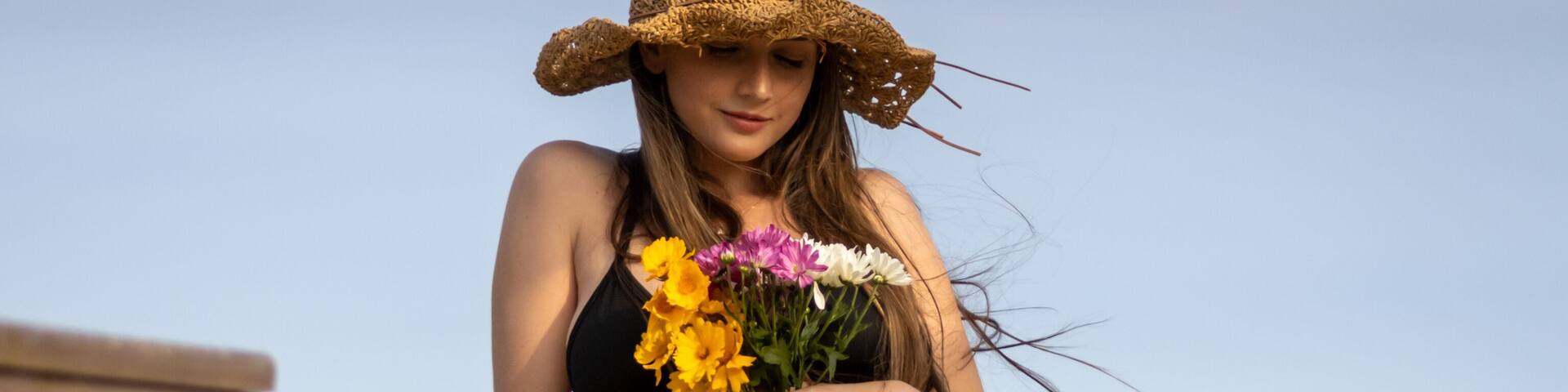 Balneário Gaivota, Santa Catarina, Brazil, November 2020. Model in front of the sea holding beautiful colorful flowers with a wavy brown hat illuminated by the sunlight.