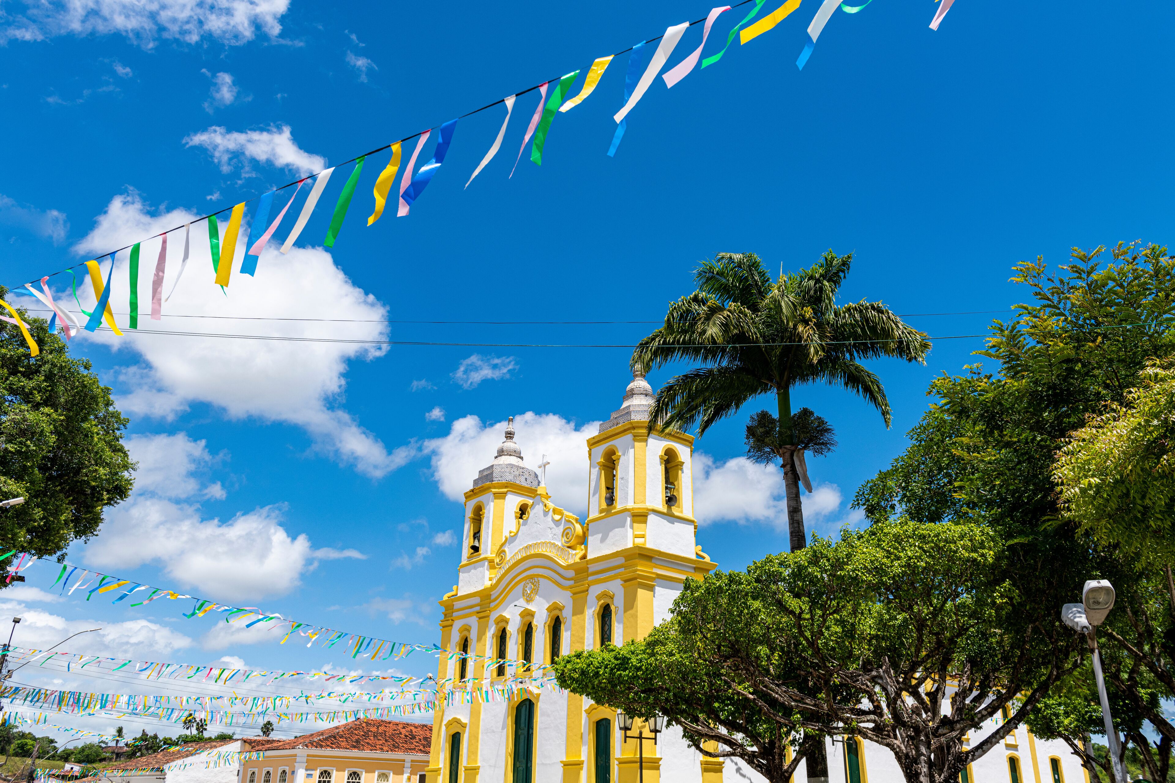 A church with yellow and white architecture, surrounded by palm trees and colorful flags.