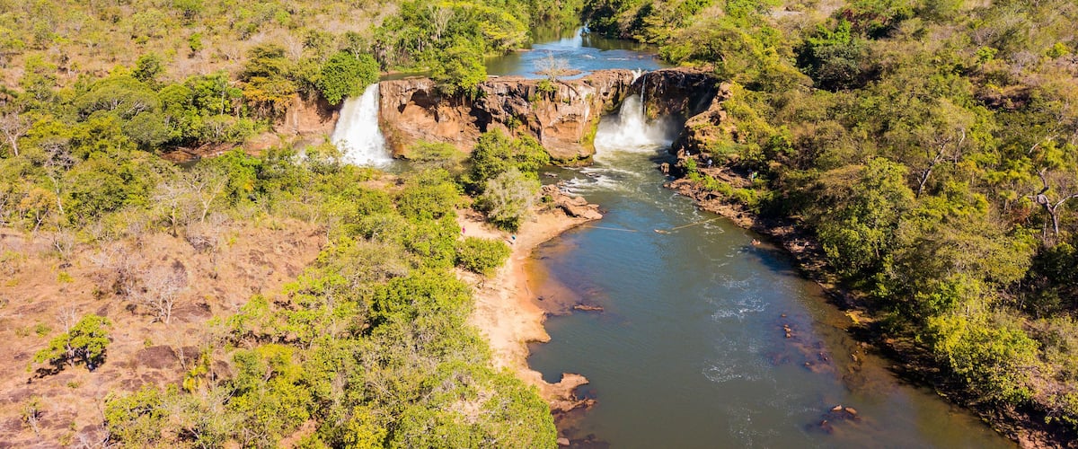A river with two waterfalls surrounded by dense green vegetation and hills in the background.