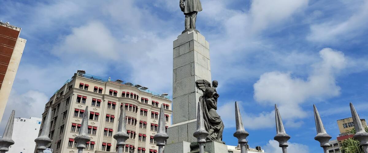 A statue of a man with outstretched arms on a tall pedestal, surrounded by ornate spires, with a historic building in the background.