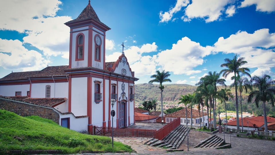 A church with a clock tower, palm trees, and a mountainous backdrop.