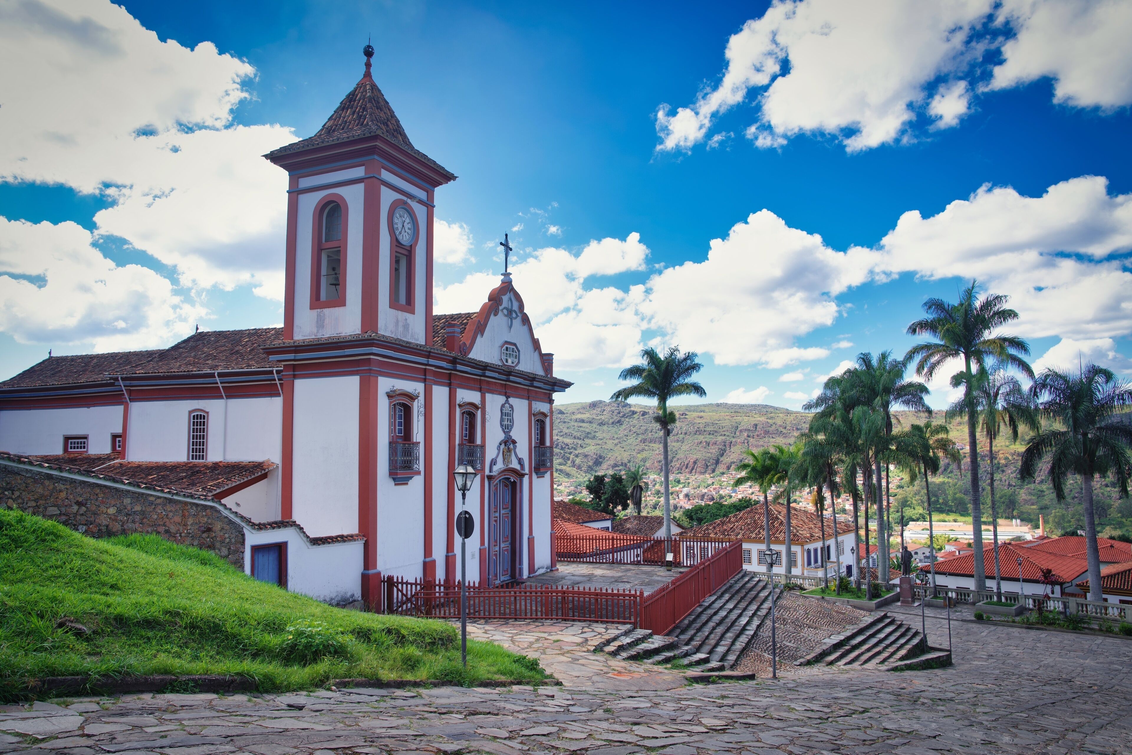A church with a clock tower, palm trees, and a mountainous backdrop.