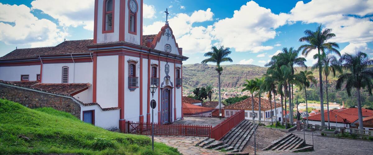 A church with a clock tower, palm trees, and a mountainous backdrop.
