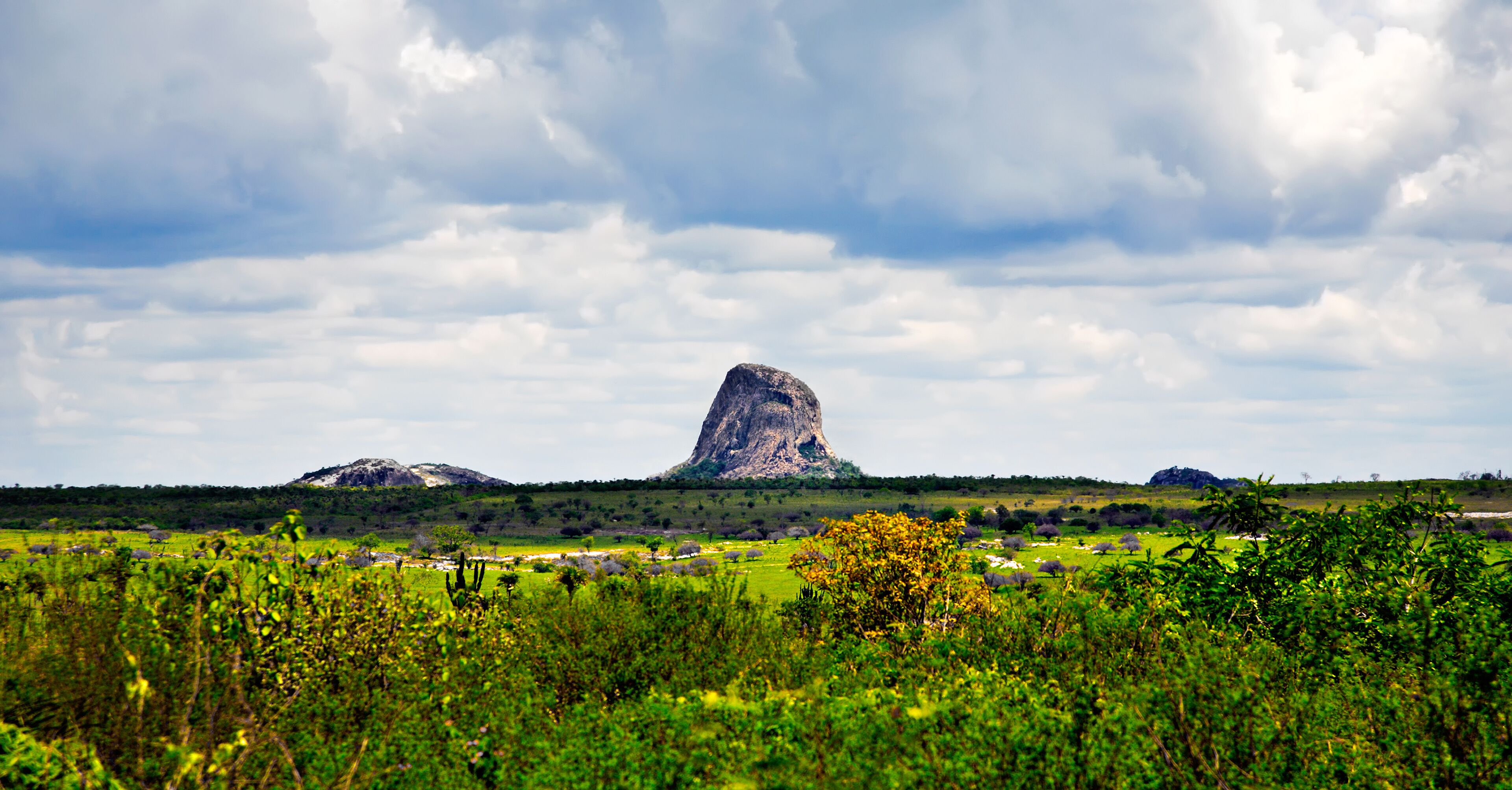 A landscape with a prominent rock formation in the center, surrounded by green fields and scattered trees.