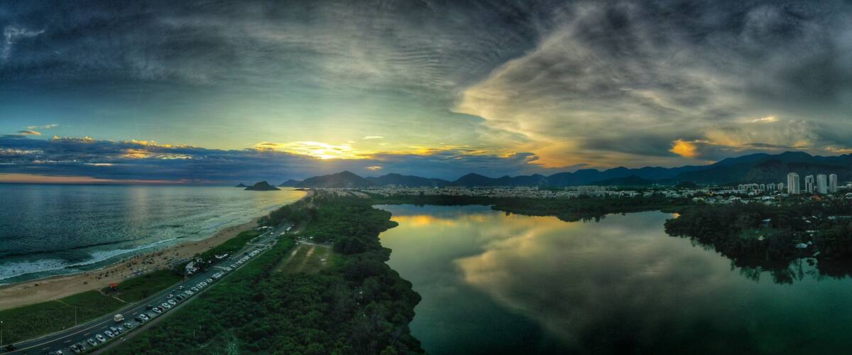 A coastal cityscape with a calm lake reflecting the sky, a beach with people, and mountains in the background.