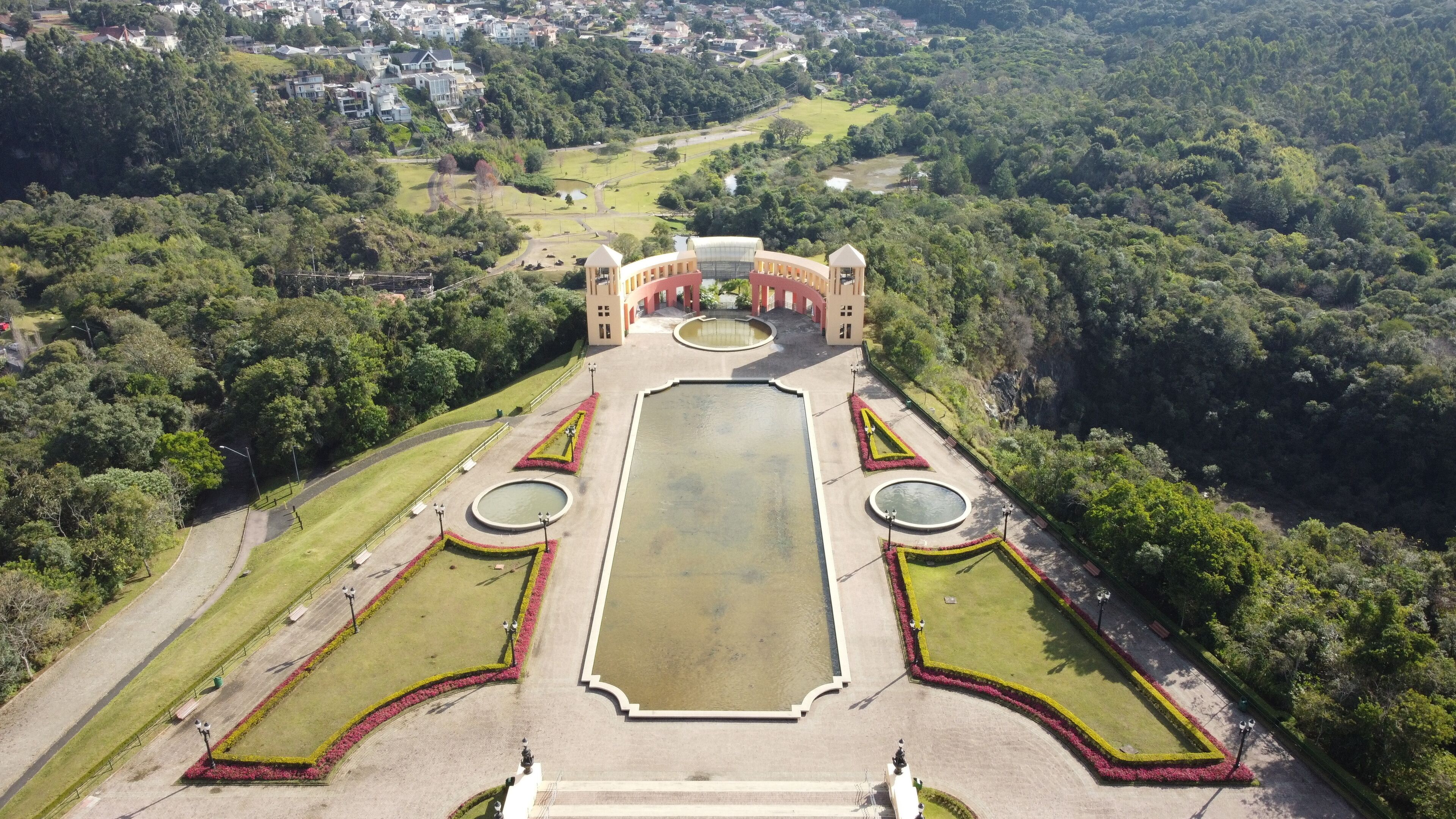An aerial view of a formal garden with a central rectangular water feature, surrounded by symmetrical flower beds and a pavilion with a dome roof.