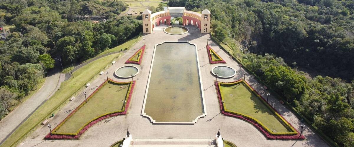 An aerial view of a formal garden with a central rectangular water feature, surrounded by symmetrical flower beds and a pavilion with a dome roof.
