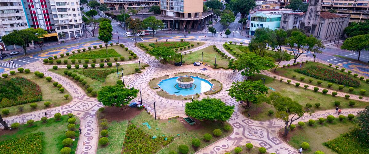 An aerial view of a landscaped public square with a central fountain, surrounded by geometrically arranged flower beds and trimmed hedges.