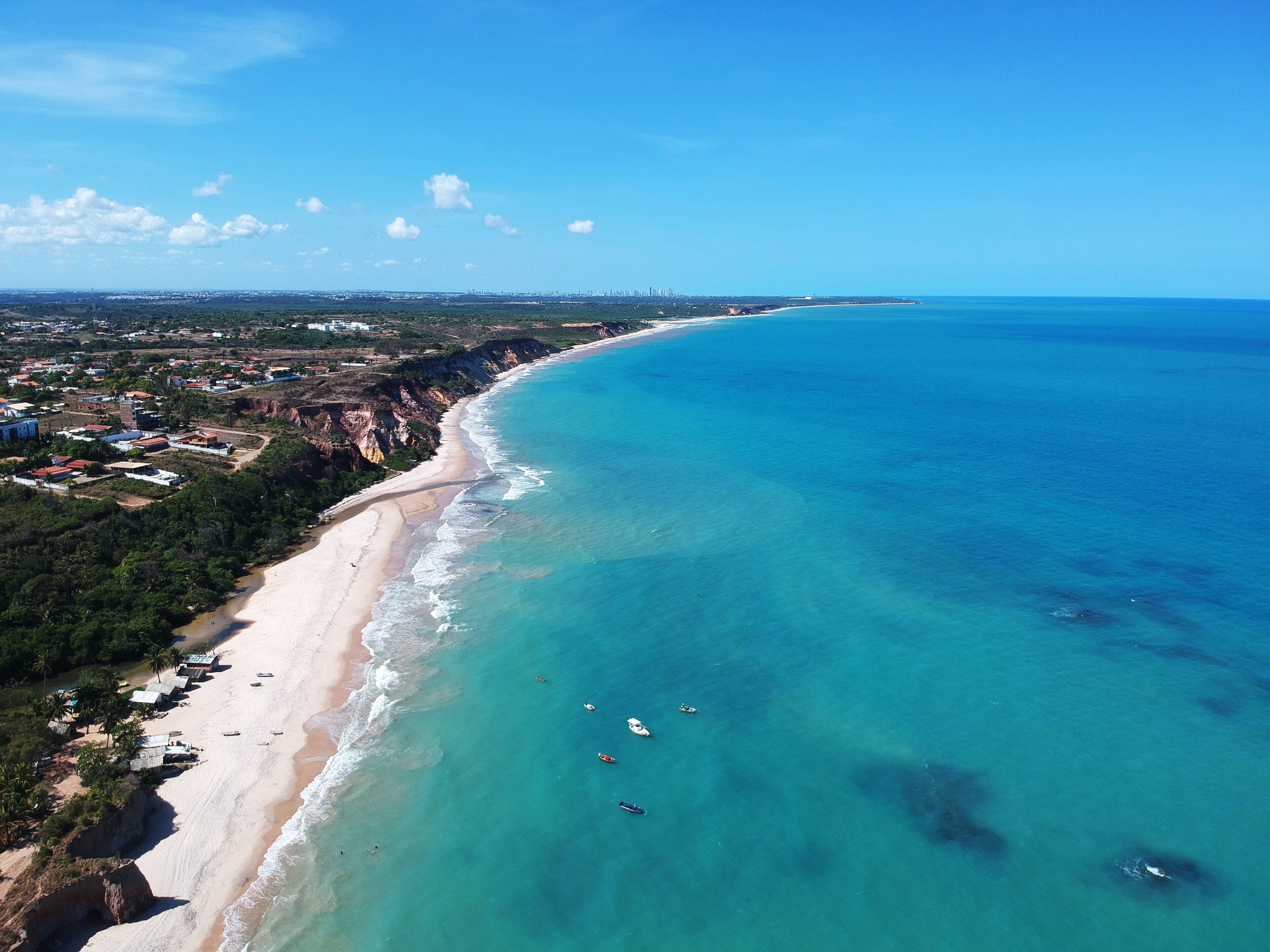 A coastal landscape with a sandy beach, clear turquoise waters, and a distant city skyline.