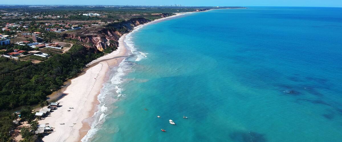 A coastal landscape with a sandy beach, clear turquoise waters, and a distant city skyline.