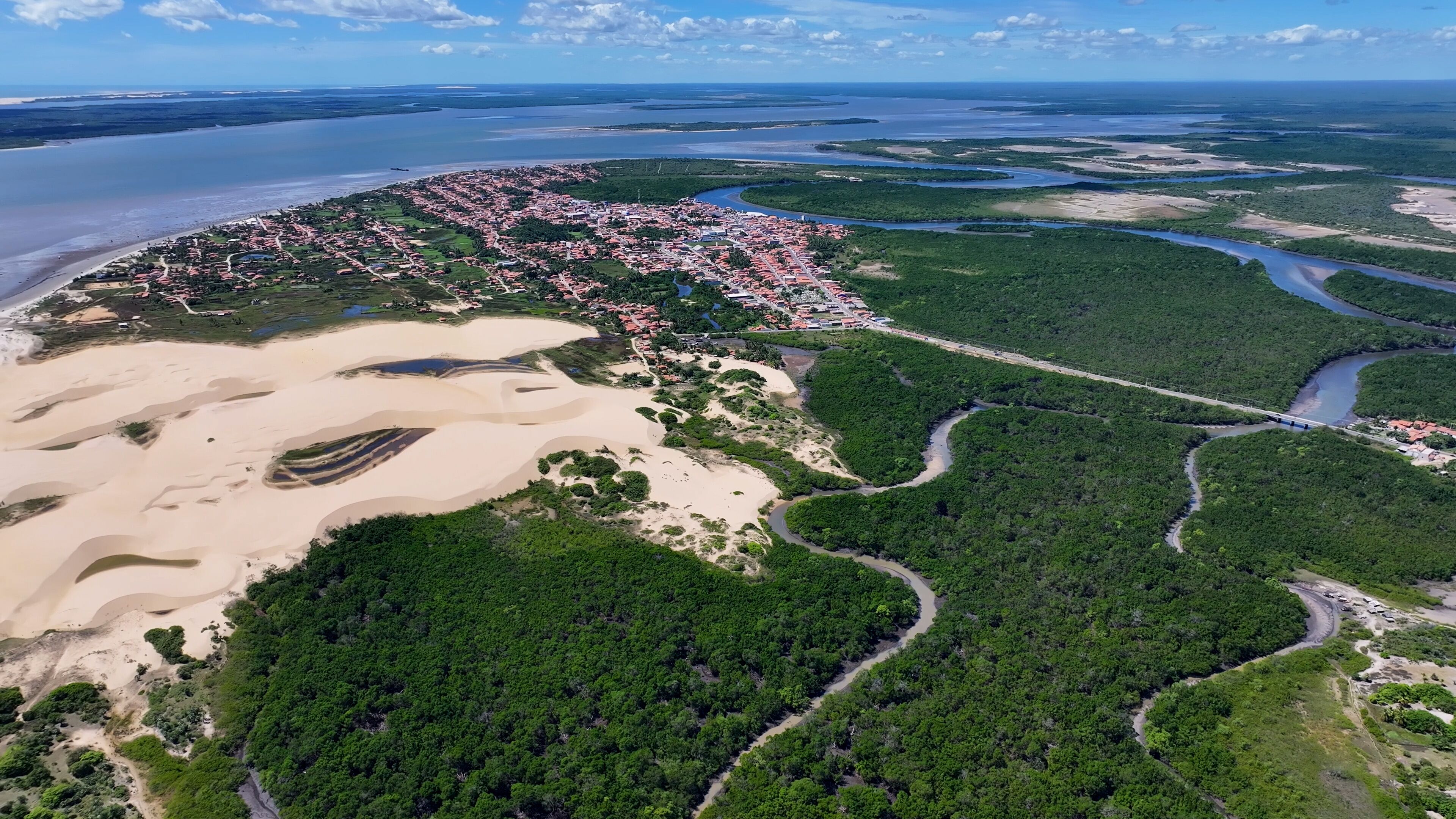 An aerial view of a coastal city with a river, dense green forest, and sandy areas.