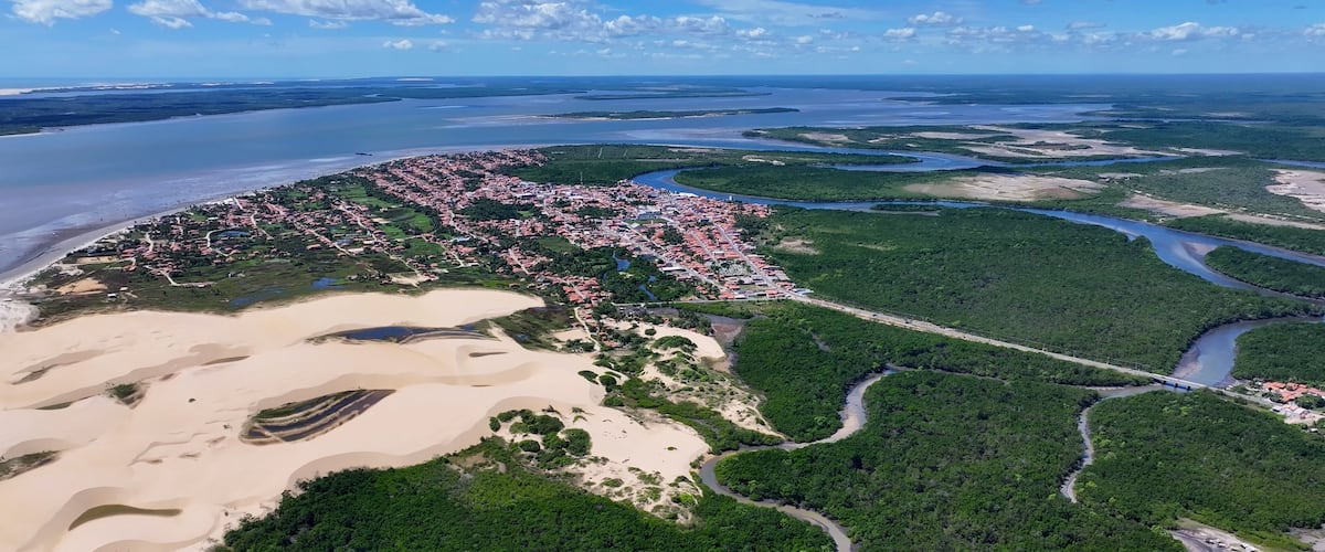 An aerial view of a coastal city with a river, dense green forest, and sandy areas.
