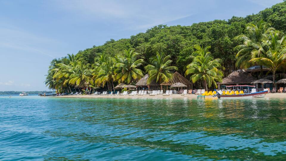 A tropical beach with palm trees, a thatched-roof hut, and a boat on the water.