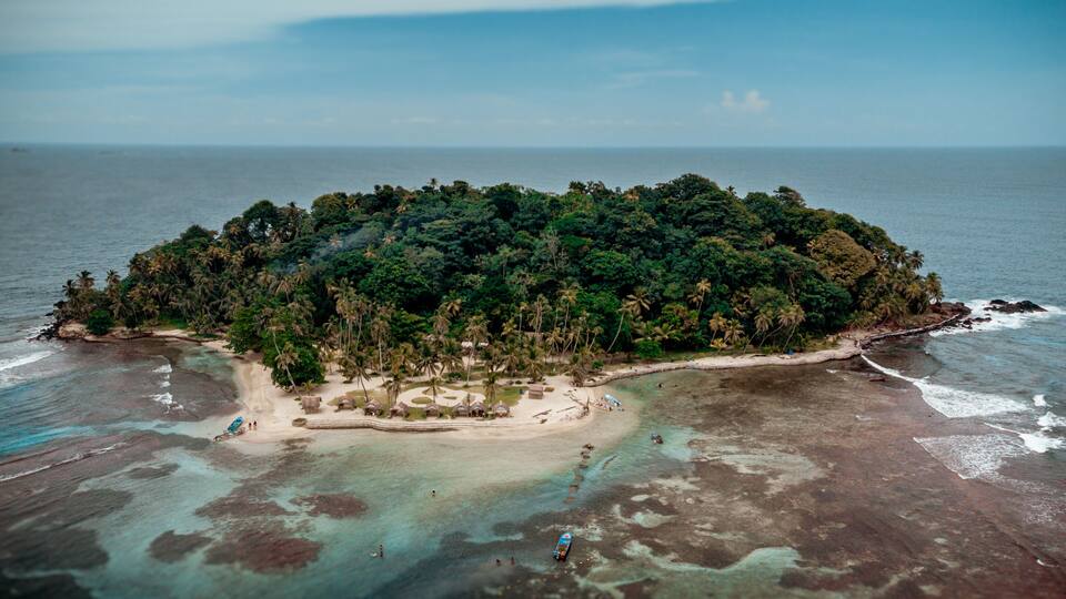 An aerial view of a tropical island with dense vegetation, clear turquoise waters, and a small beach area with thatched-roof huts.