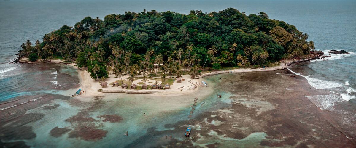 An aerial view of a tropical island with dense vegetation, clear turquoise waters, and a small beach area with thatched-roof huts.
