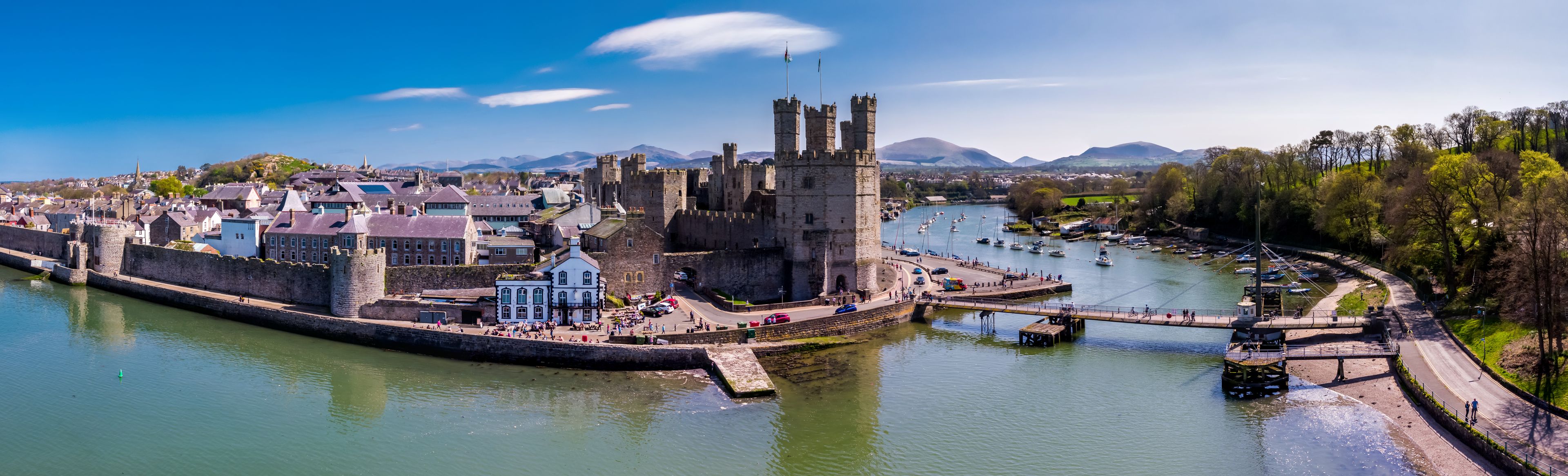 A historic town with a prominent castle, a river with boats, and a mountainous backdrop.