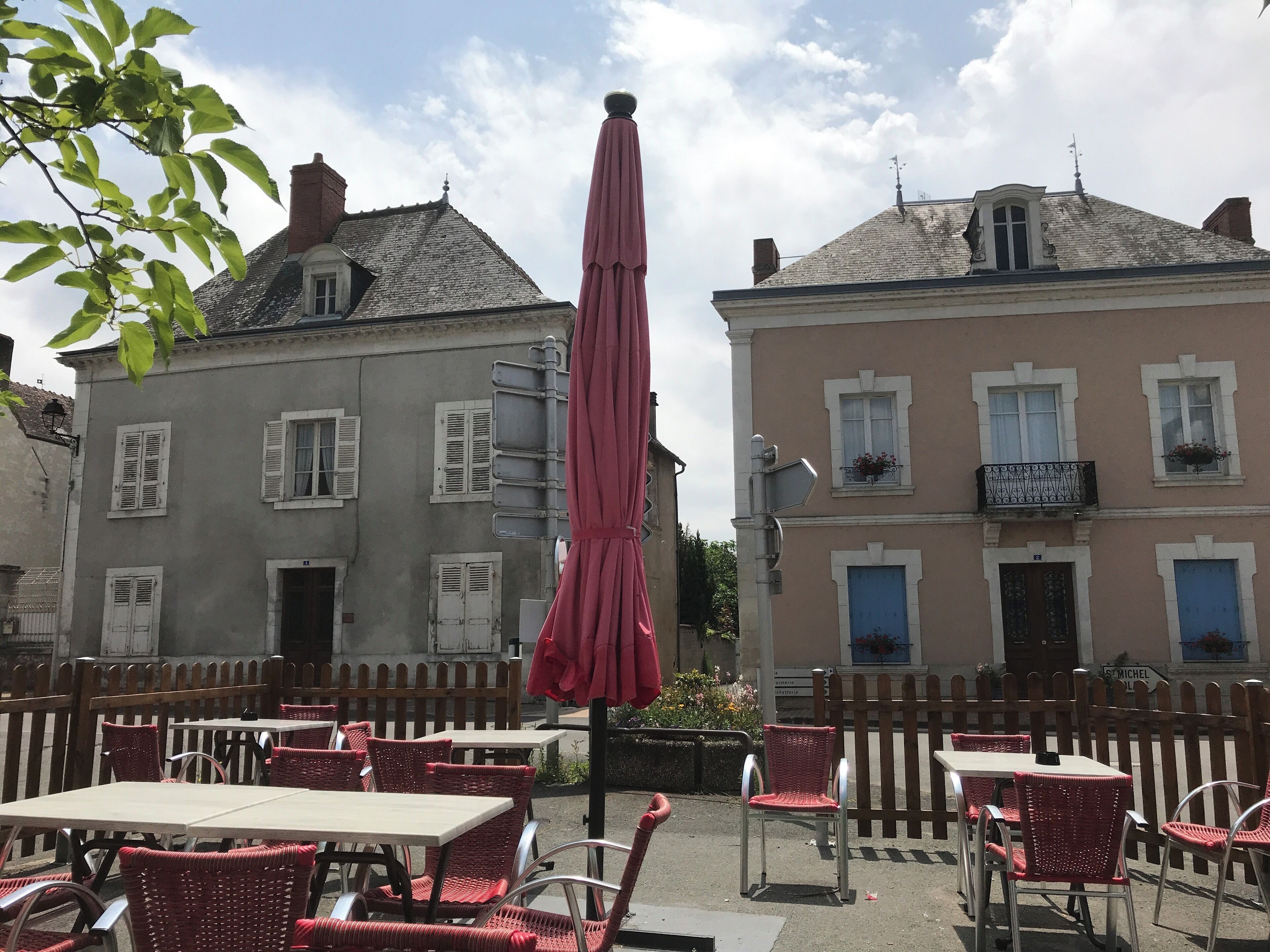 An outdoor café with red wicker chairs and tables, a pink umbrella, and buildings with white shutters in the background.