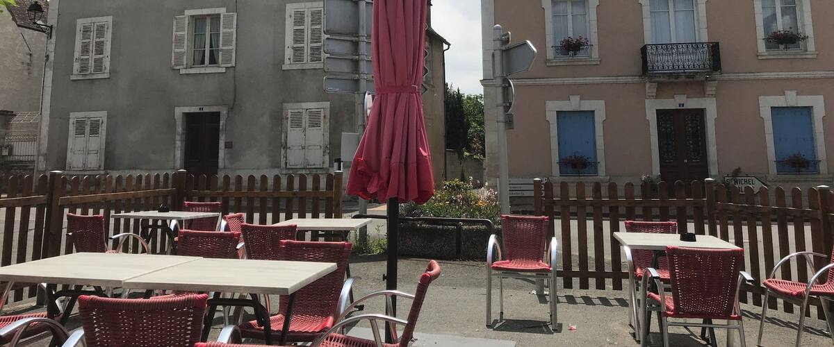 An outdoor café with red wicker chairs and tables, a pink umbrella, and buildings with white shutters in the background.