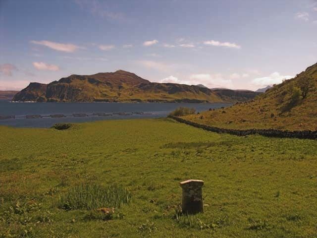 A grassy field with a stone marker, a body of water, and hills in the background.