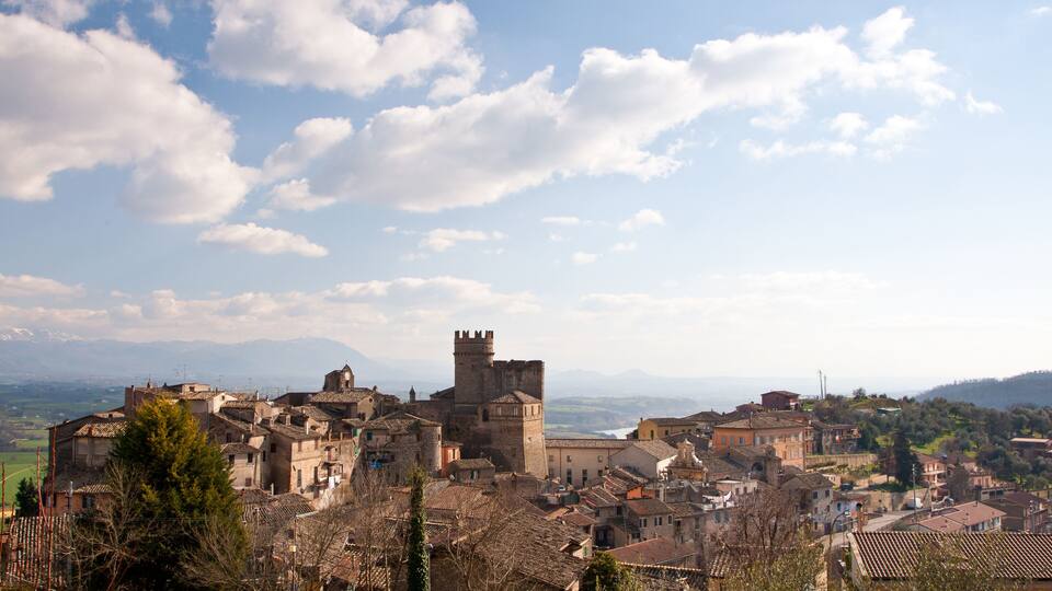 A historic town with a prominent castle, terracotta-roofed buildings, and a clear blue sky with scattered clouds.