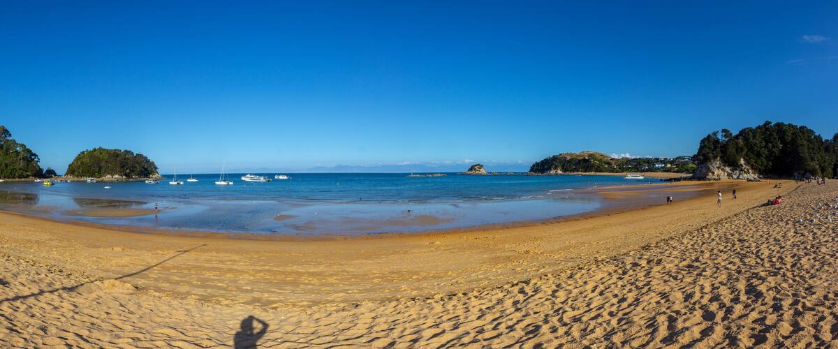 A sandy beach with clear blue water, boats, and a forested island in the distance.