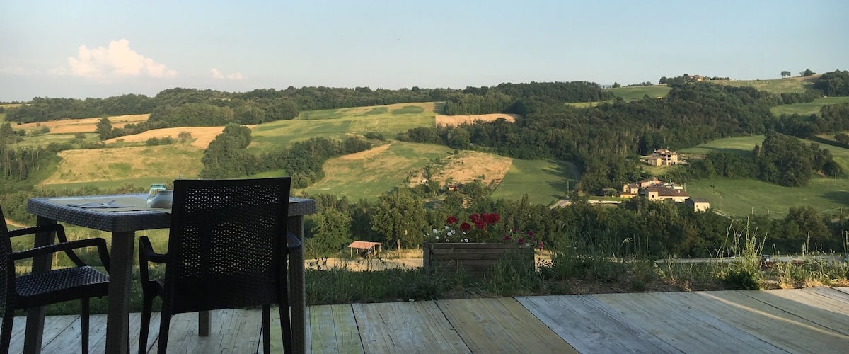 A wooden deck with a table and chairs overlooking rolling hills and a distant village.