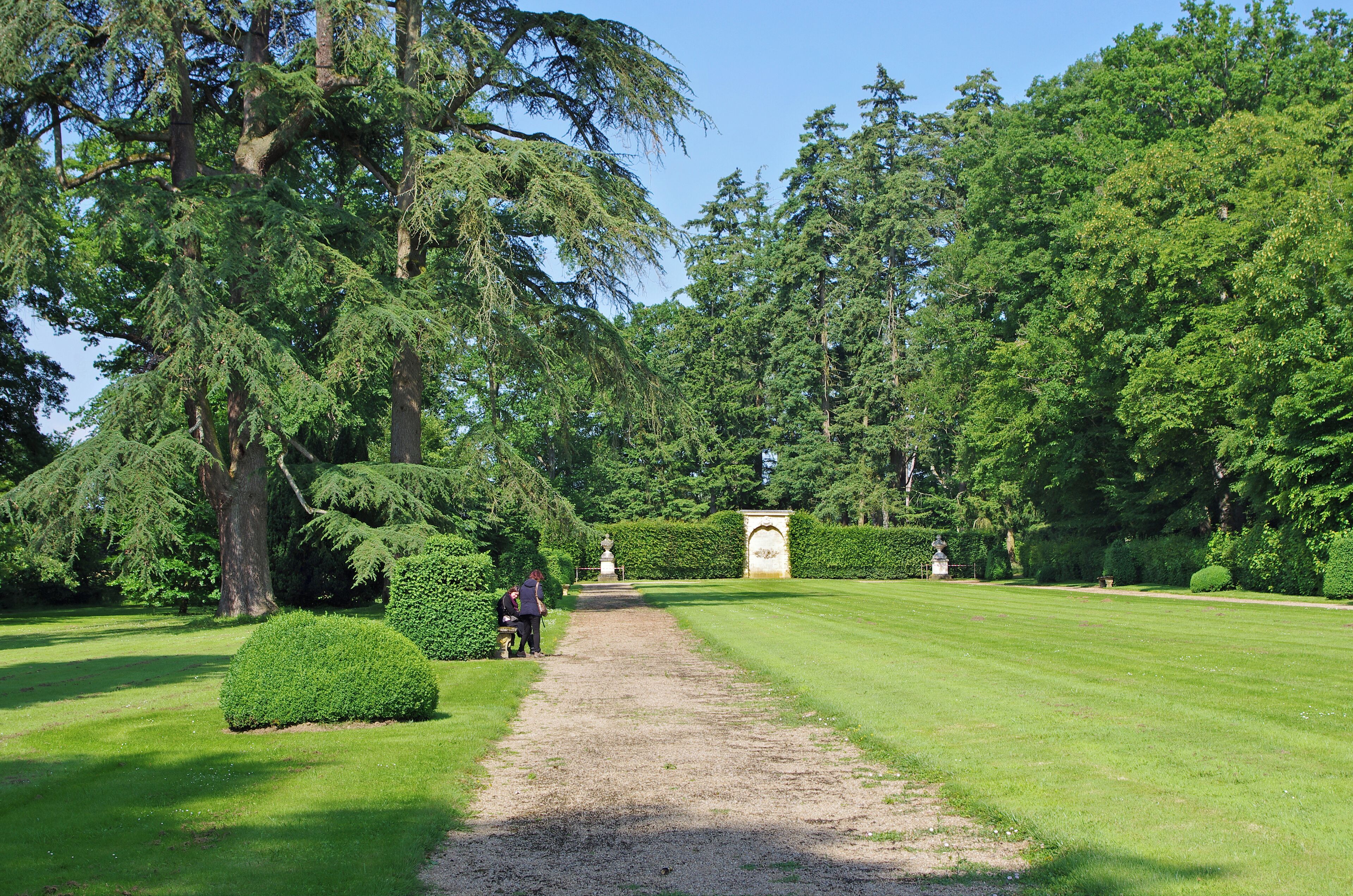 A well-maintained garden with a gravel path, neatly trimmed hedges, and tall trees.