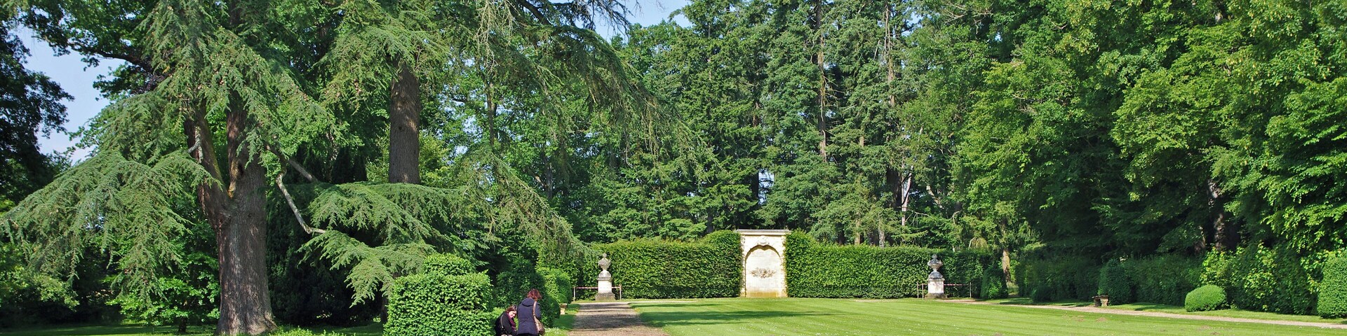 A well-maintained garden with a gravel path, neatly trimmed hedges, and tall trees.