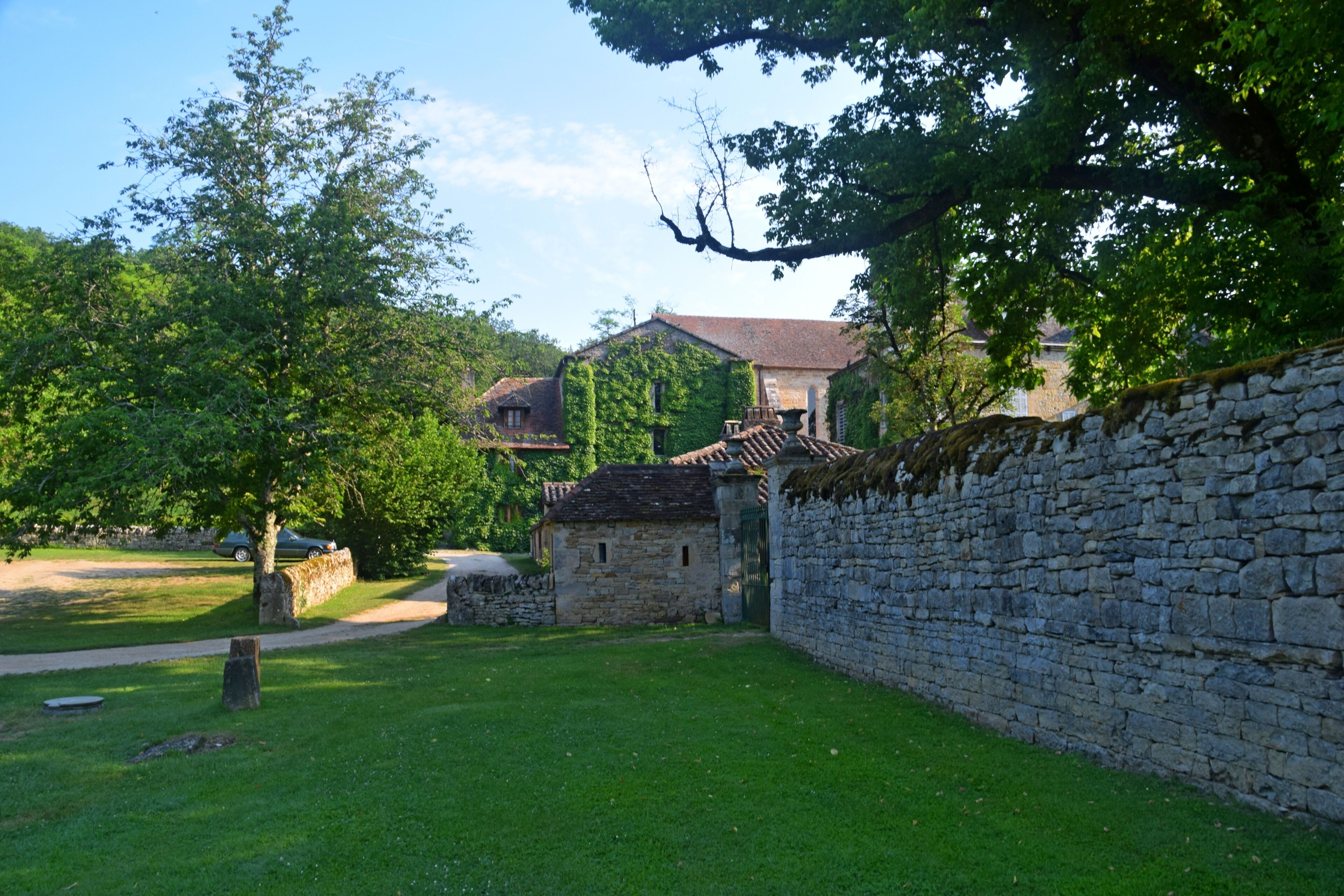 A stone wall, a stone building with a tiled roof, and a tree in a grassy area.