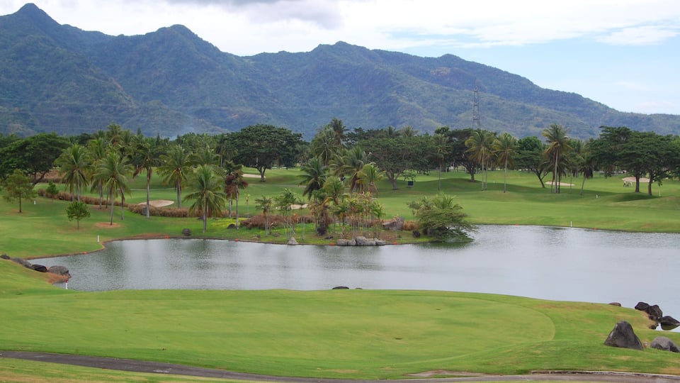 A golf course with a pond, palm trees, and mountains in the background.