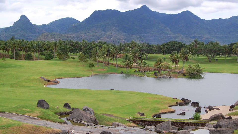 A golf course with a pond, palm trees, and mountains in the background.