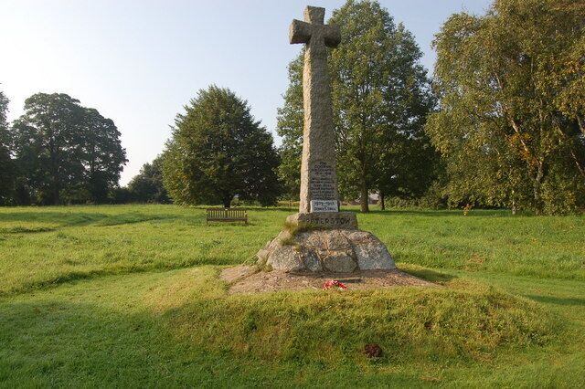 A stone cross monument in a grassy field with trees in the background.
