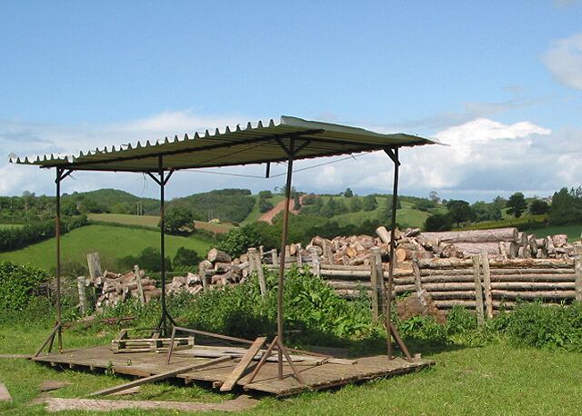 A rustic wooden structure with a metal roof, surrounded by a pile of logs and wooden planks in a grassy field with rolling hills in the background.