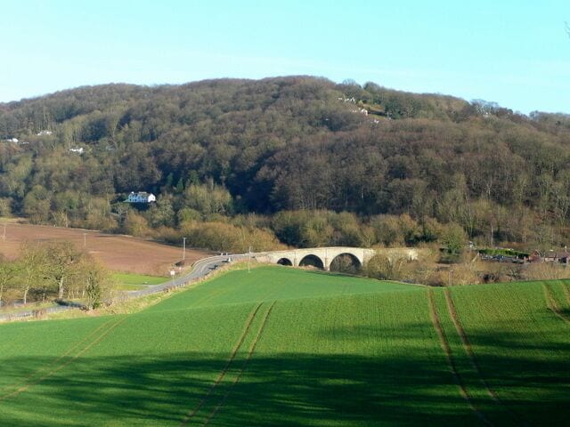 Un puente sobre un río, campos verdes y una colina boscosa.