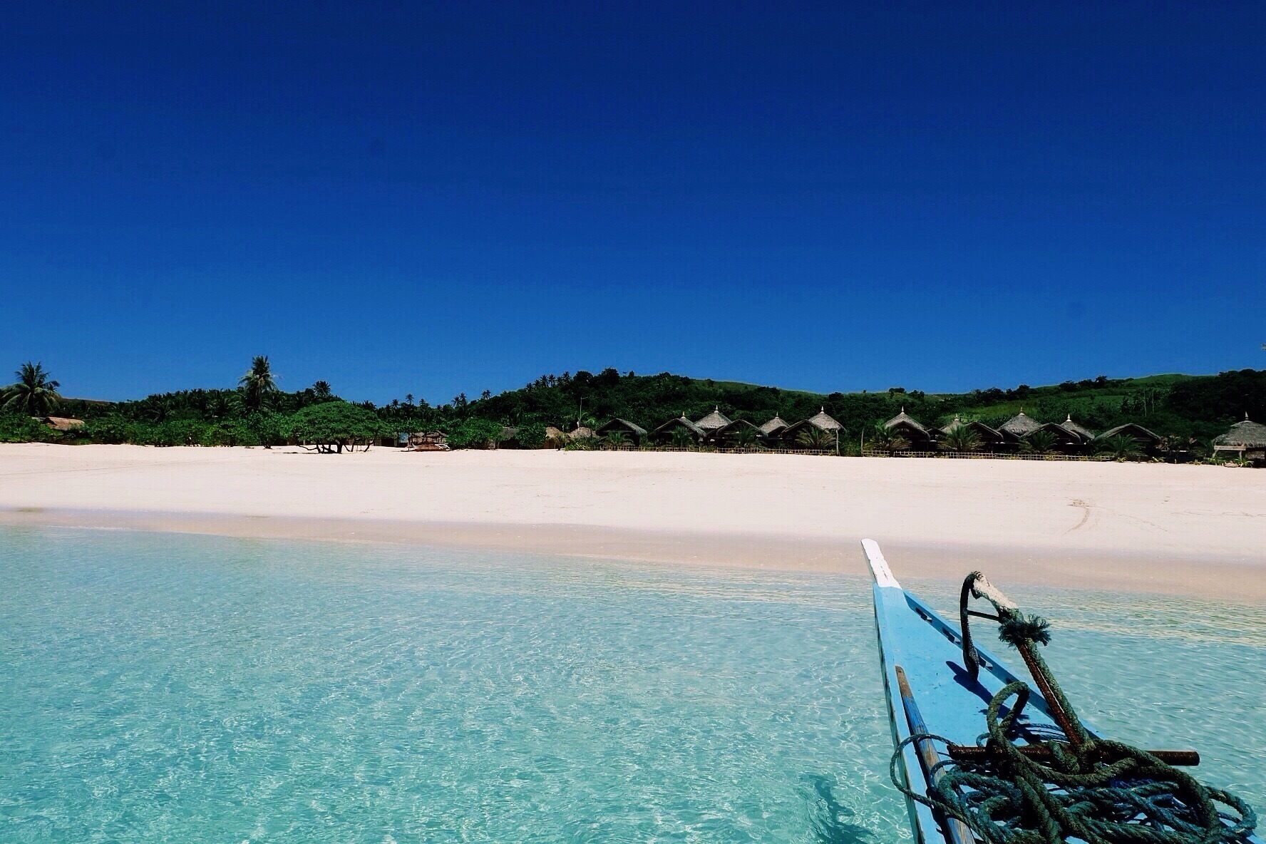 A beach with clear turquoise water, white sand, and thatched-roof huts in the background.