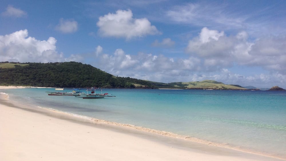 Une plage à l’eau turquoise et limpide, un rivage sablonneux et plusieurs bateaux ancrés près du bord de l’eau.