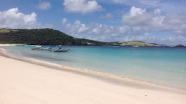Uma praia com águas turquesa cristalinas, areal arenoso e vários barcos ancorados perto da beira-mar.
