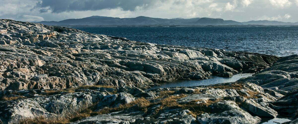 A rocky coastline with a calm sea and distant mountains under a cloudy sky.