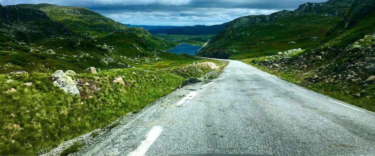 A winding mountain road flanked by lush green hills and a lake under a partly cloudy sky.