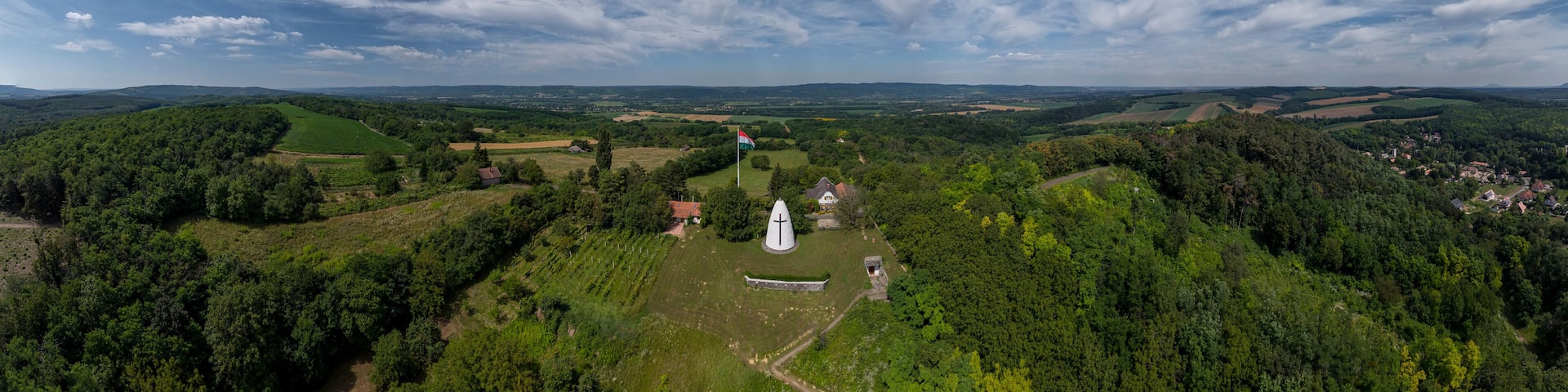 A panoramic view of a lush green valley with rolling hills, a white church, and a small village in the distance under a partly cloudy sky.