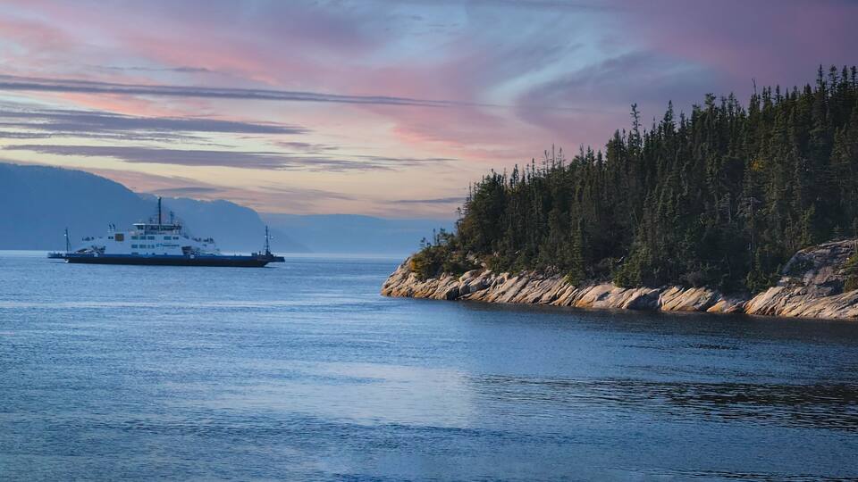 A ferry on a calm body of water with a forested coastline and mountains in the background.