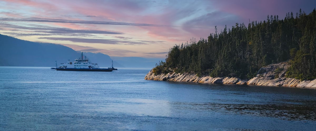 A ferry on a calm body of water with a forested coastline and mountains in the background.