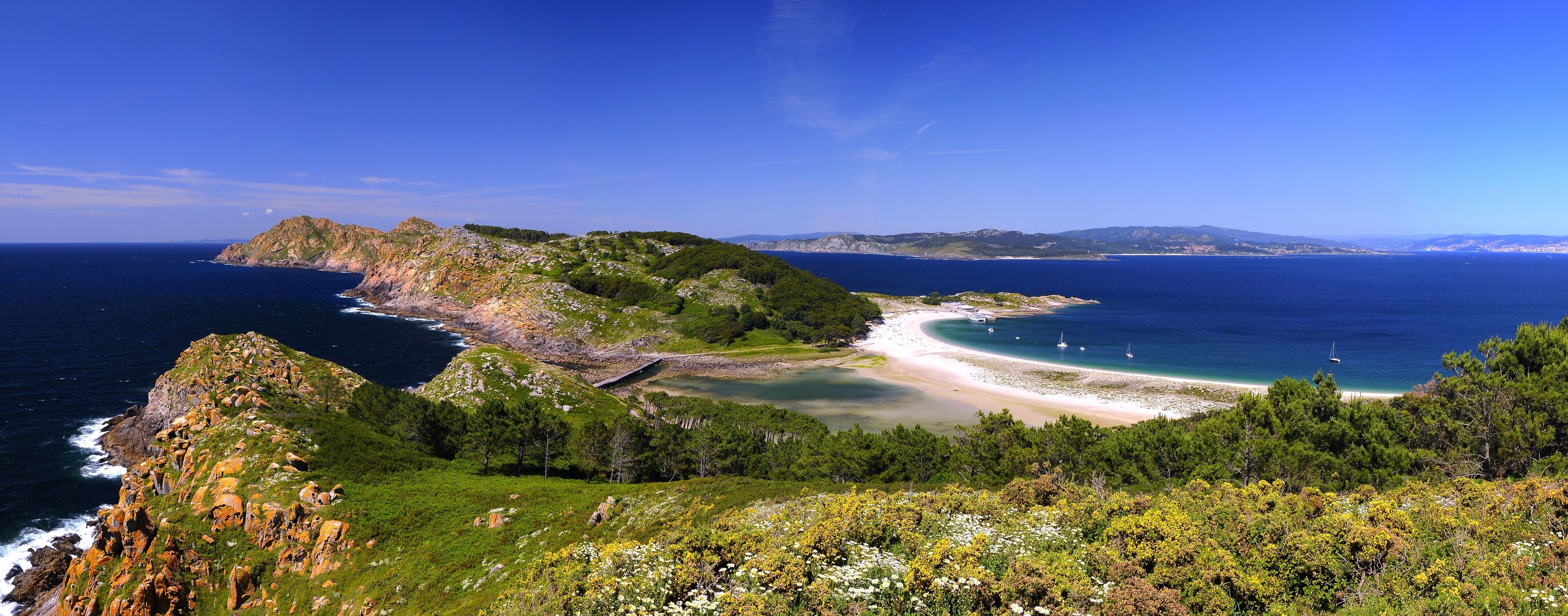 A coastal landscape with a rocky peninsula, a sandy beach, and a clear blue sea.