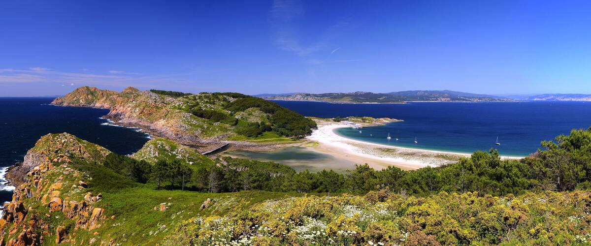 A coastal landscape with a rocky peninsula, a sandy beach, and a clear blue sea.