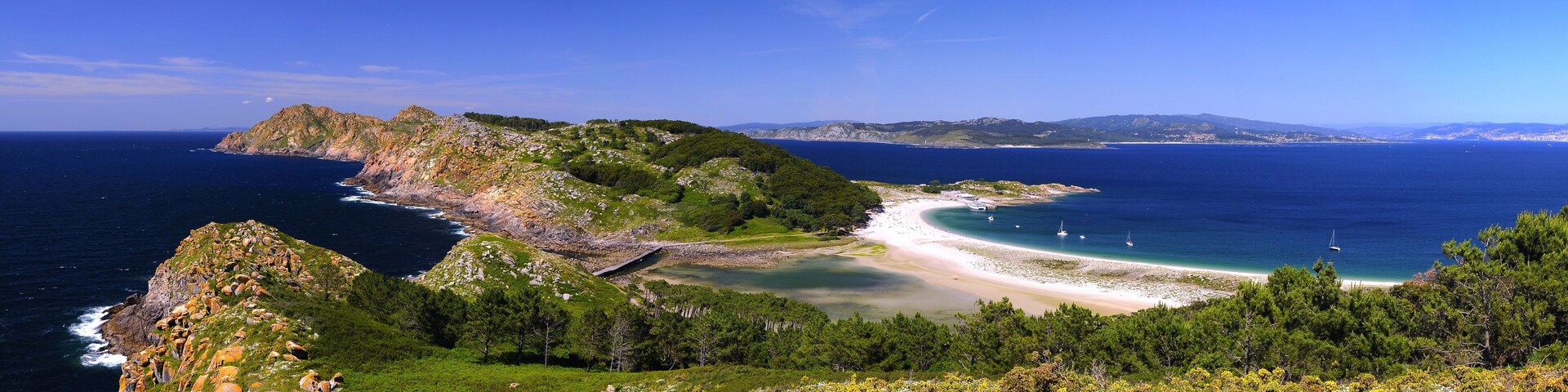 A coastal landscape with a rocky peninsula, a sandy beach, and a clear blue sea.